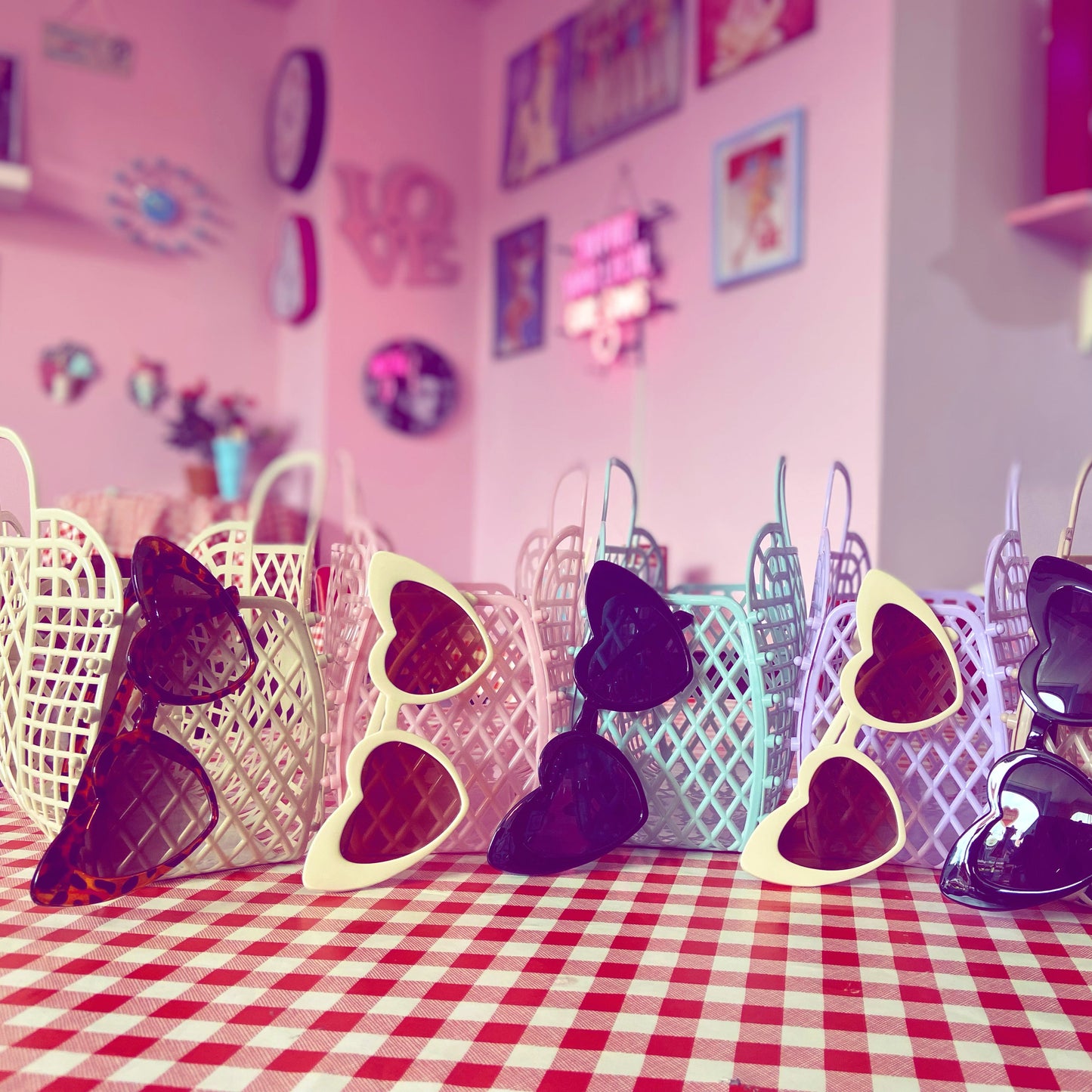Heart-shaped sunglasses in decorative baskets on a checkered tablecloth with a pink wall background.