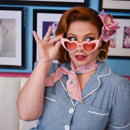 Woman in a vintage-style dress with heart-shaped sunglasses and a flower in her hair, posing in front of framed pictures.